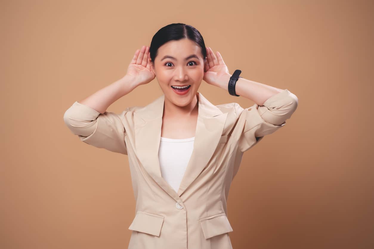 Happy woman holding her hands to her ears on a tan background.